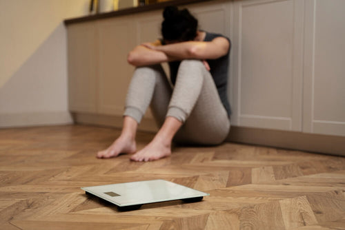 A woman sitting on the floor beside a bathroom scale looking frustrated representing the emotional toll of chasing scale weight without understanding the difference between losing weight and losing fat.
