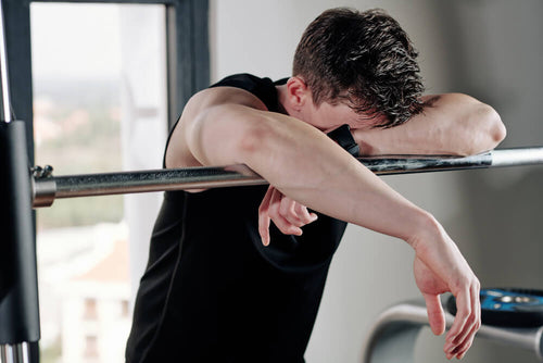 A fit man's torso visible leaning over a barbell in a gym looking fatigued and really sore from working out after a heavy lifting session.