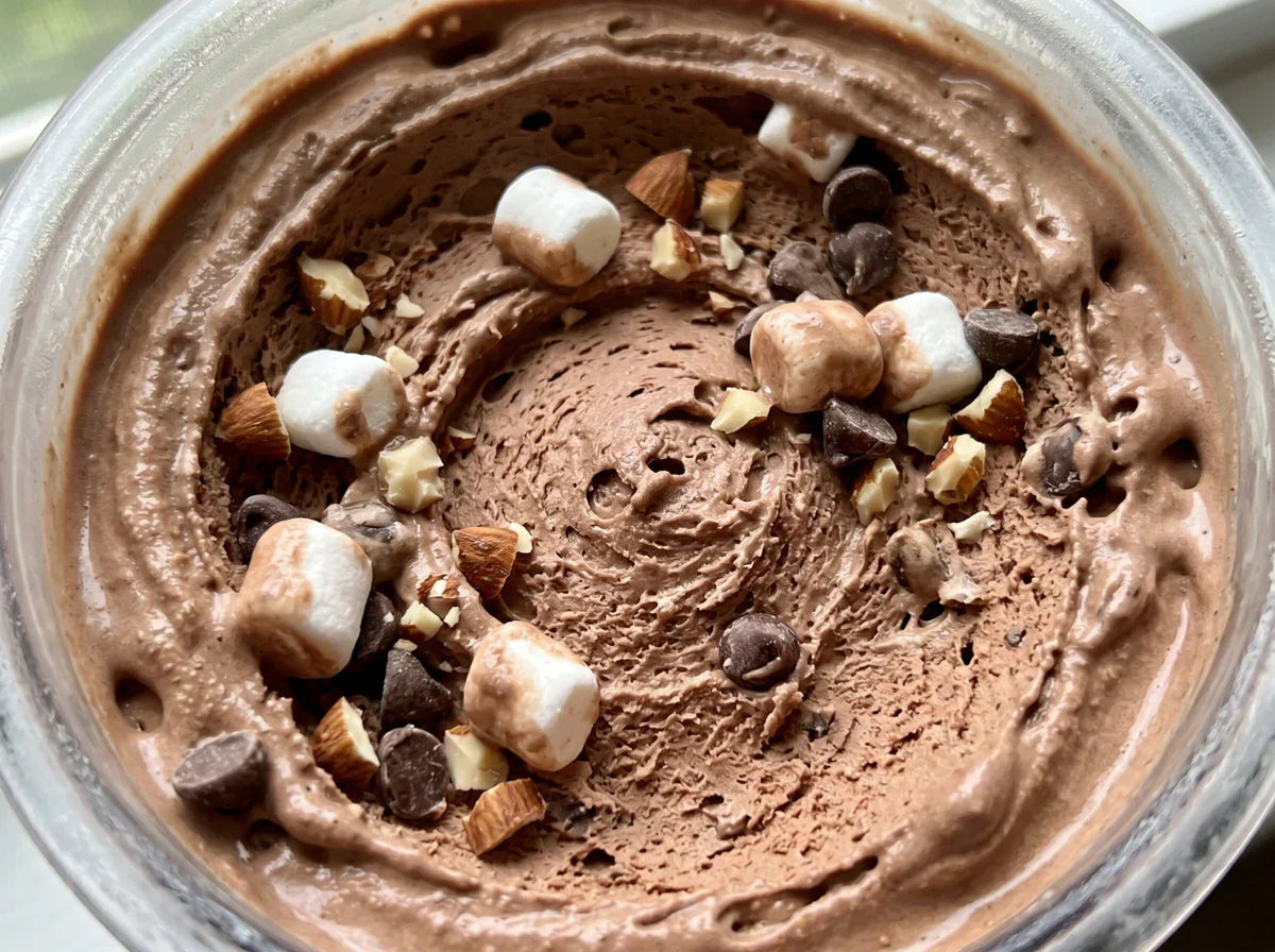 Close-up of Rocky Road protein ice cream in a Ninja Creami pint, with marshmallows, chopped almonds, and chocolate chips mixed into creamy chocolate base.