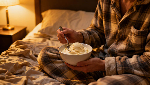 A person's hand scooping creamy vanilla ice cream from a bowl while sitting in bed under warm blankets representing one of the most enjoyable bedtime protein snacks for overnight muscle recovery.