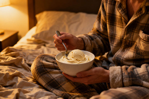 A person's hand scooping creamy vanilla ice cream from a bowl while sitting in bed under warm blankets representing one of the most enjoyable bedtime protein snacks for overnight muscle recovery.