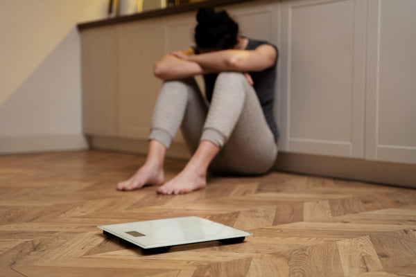 A woman sitting on the floor beside a bathroom scale looking frustrated representing the emotional toll of chasing scale weight without understanding the difference between losing weight and losing fat.