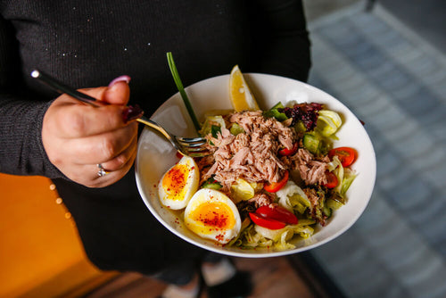 A person's hands visible eating a bowl of tuna salad with hard boiled eggs and vegetables representing a practical GLP-1 meal plan built around high protein foods that are easy to eat in small portions.