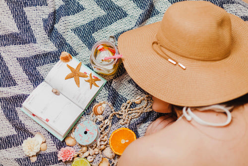 A flat lay of a straw hat sunglasses a journal and seashells on a beach towel representing the relaxed intentional lifestyle behind how to build a summer routine that actually fits your life.