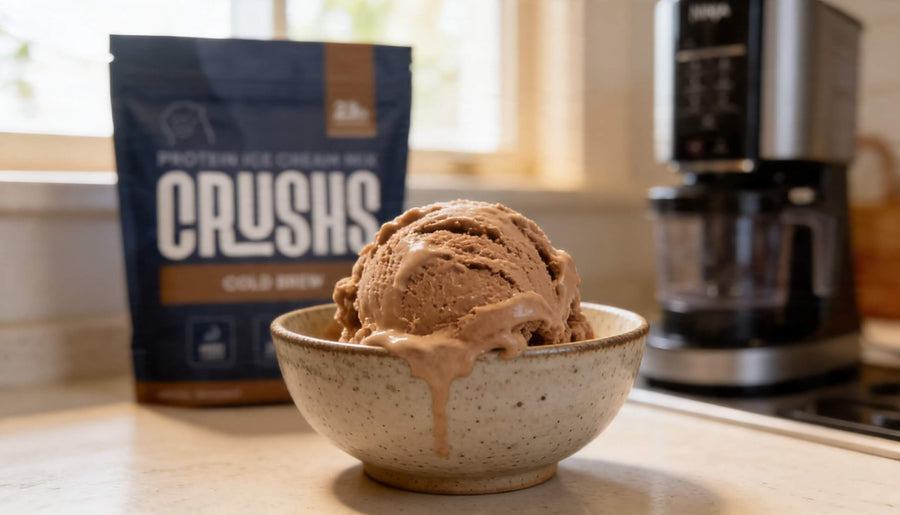 A creamy scoop of cold brew protein ice cream in a speckled ceramic bowl with a CRUSHS Cold Brew Protein Ice Cream Mix bag and a Ninja Creami machine visible in the background showing how to make Ninja Creami creamy with the right base.