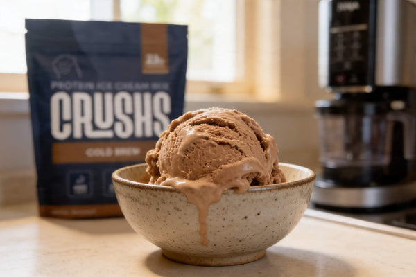 A creamy scoop of cold brew protein ice cream in a speckled ceramic bowl with a CRUSHS Cold Brew Protein Ice Cream Mix bag and a Ninja Creami machine visible in the background showing how to make Ninja Creami creamy with the right base.