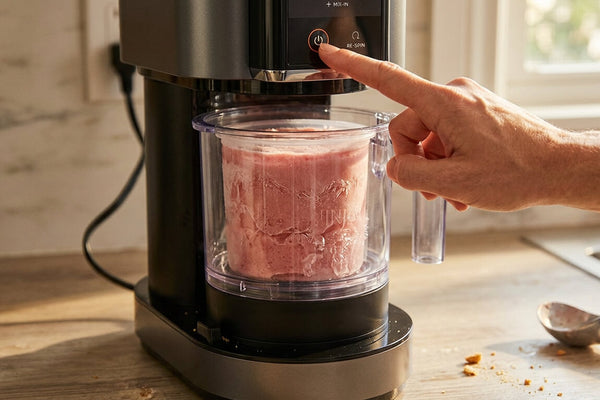 A hand pressing the start button on a Ninja Creami machine with a frozen strawberry pint visible inside representing the moment when a ninja creami too hard to spin problem stops the machine before processing begins.