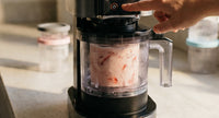 A hand pressing a button on a Ninja Creami machine with a strawberry pint visible inside representing the hands-on ninja creami troubleshooting process for a machine that is struggling to process correctly.