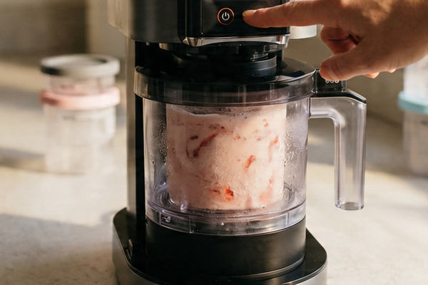 A hand pressing a button on a Ninja Creami machine with a strawberry pint visible inside representing the hands-on ninja creami troubleshooting process for a machine that is struggling to process correctly.