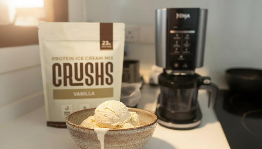 A Ninja Creami machine on a kitchen counter beside a CRUSHS Protein Ice Cream Mix bag and a freshly spun scoop of protein ice cream recipe ninja creami style, warm natural light.