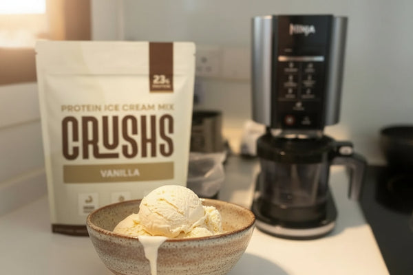 A Ninja Creami machine on a kitchen counter beside a CRUSHS Protein Ice Cream Mix bag and a freshly spun scoop of protein ice cream recipe ninja creami style, warm natural light.