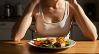 A woman's torso visible sitting in front of an untouched plate of grilled chicken and vegetables with her hands on her head representing the frustration of not being able to hit protein on GLP-1 due to appetite suppression.