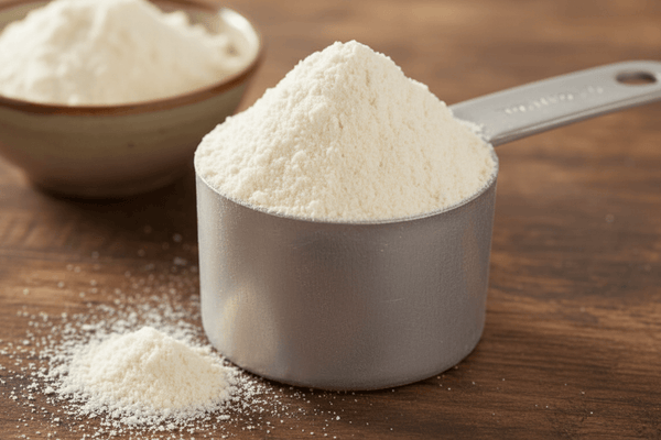 A metal scoop heaped with fine white milk protein isolate powder on a wooden surface, with loose powder dusted around it and a ceramic bowl blurred in the background.