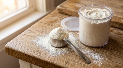 A metal scoop of fine white protein powder beside a Ninja Creami pint container on a wooden surface showing the process of choosing what protein powder to use Ninja Creami for a creamy consistent base.