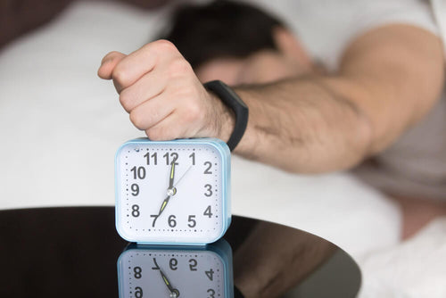 Person sleeping in bed while reaching to turn off an alarm clock, representing tiredness after 8 hours of sleep.