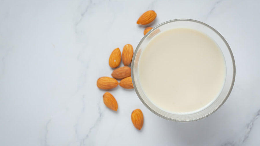 Glass of almond milk viewed from above with raw almonds scattered on a white marble surface.