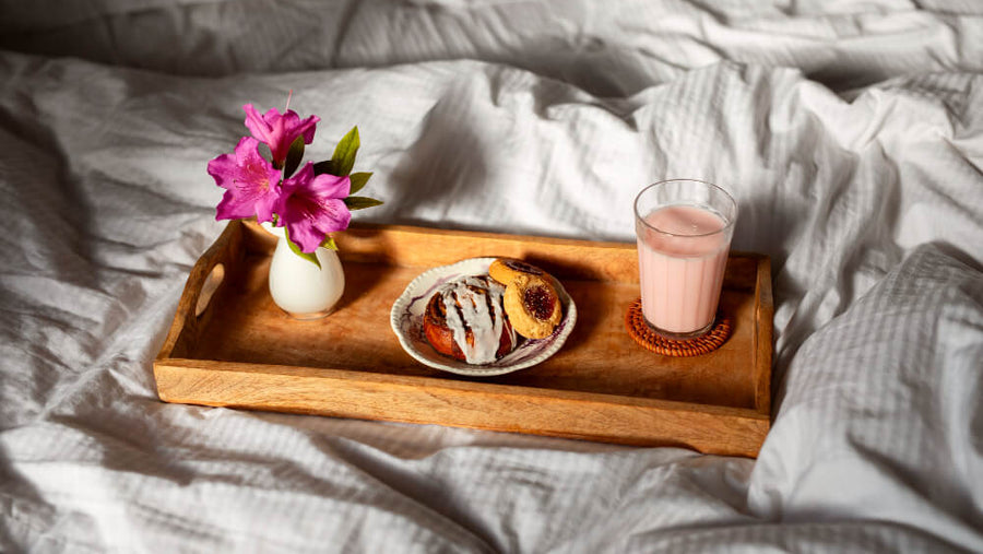A wooden breakfast tray with pastries, a pink drink, and fresh flowers on white linen bedding for Mother's Day morning.