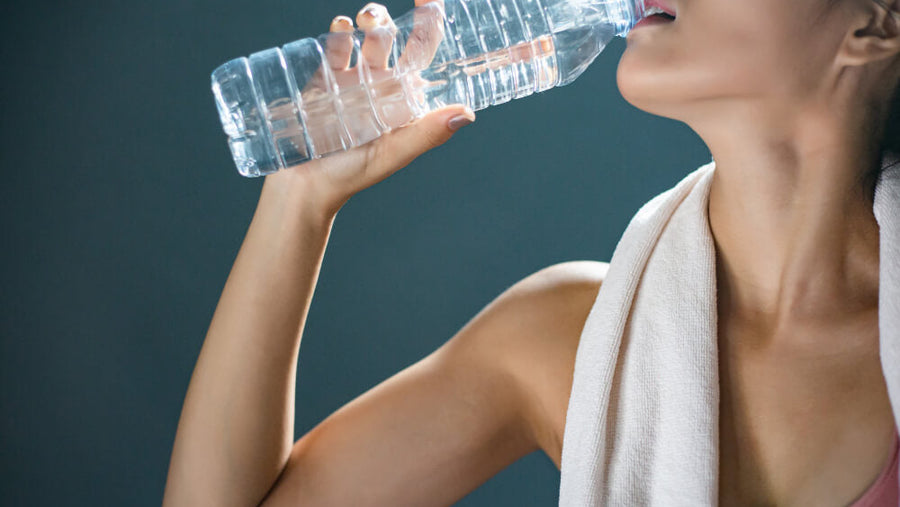 A woman's torso visible drinking water from a bottle with a towel over her shoulder representing the hydration habits essential to how to get ready for summer body through consistent daily basics.