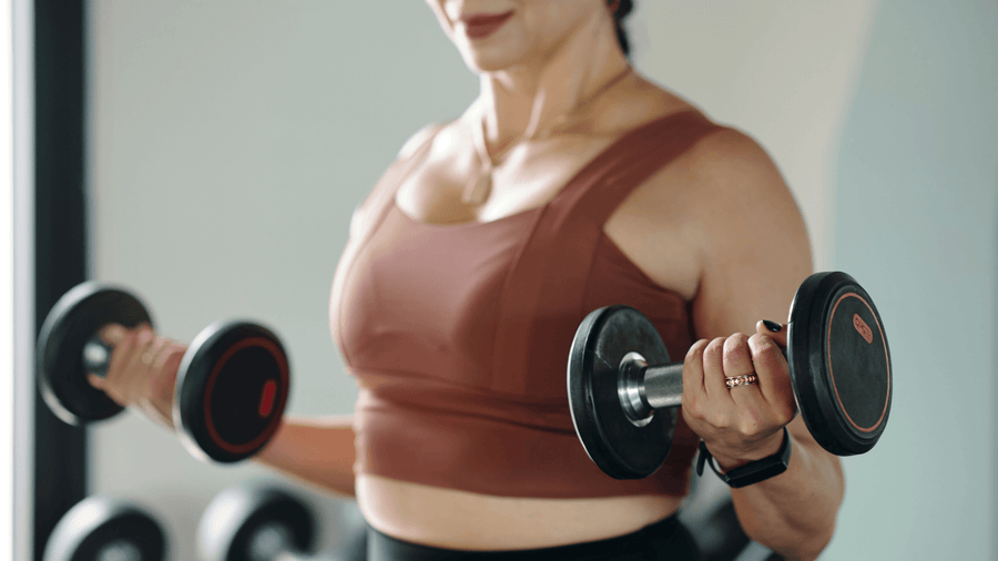 A fit woman's torso and arms visible curling a dumbbell in gym clothes representing the strength training component of how to get ready for summer body by building and preserving lean muscle.