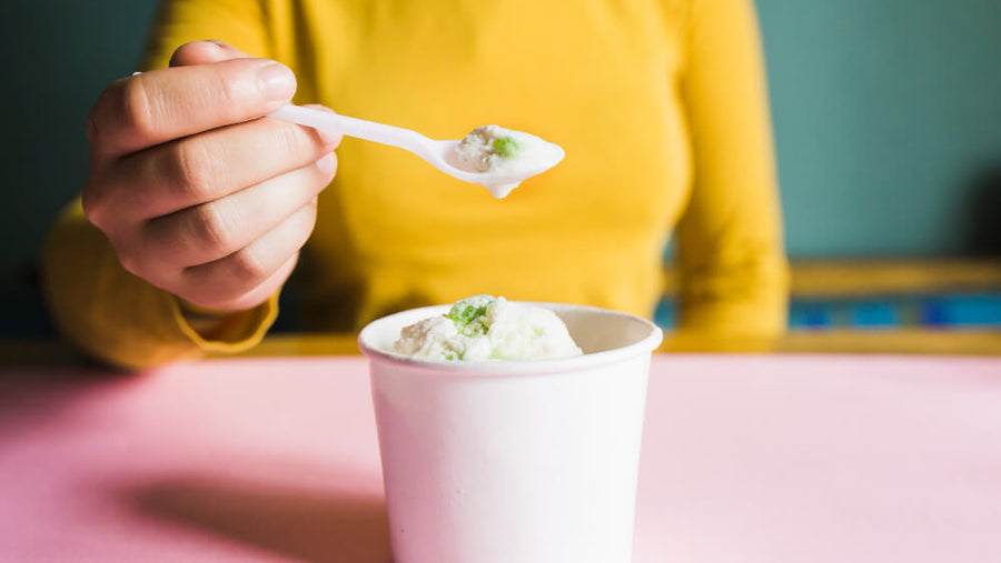 A woman's torso visible in a yellow sweater eating protein ice cream from a paper cup with a spoon showing that is protein ice cream healthy is a yes when the protein per serving actually moves the needle.