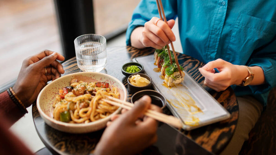 Two people sharing sushi and noodles at a restaurant table with chopsticks and dipping sauce.