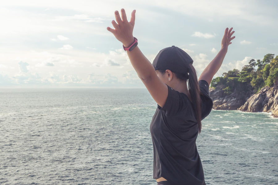 Woman in athletic wear standing by the ocean with arms raised, feeling free and energized