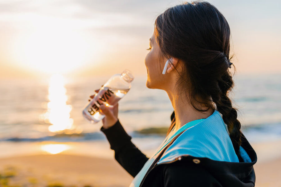 A woman's profile visible drinking water from a bottle outdoors at sunset with earphones in representing the hydration habits essential to how to feel good this summer especially in the heat.