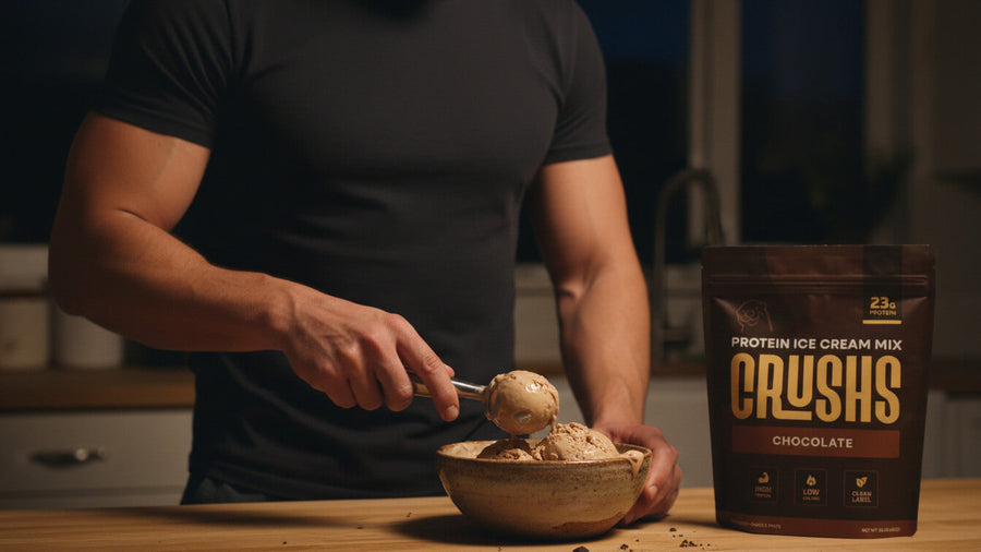 A muscular man's torso and arms visible standing at a kitchen counter with a CRUSHS Protein Ice Cream Mix bag beside him showing how casein vs whey protein applies to real nighttime nutrition habits.