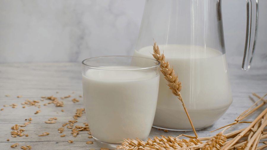 Oat milk being poured into a glass next to a wheat stalk as a clean healthy coffee add-in alternative to sugary creamer.