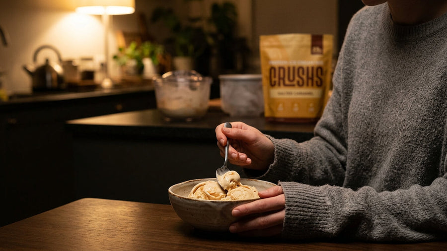 A person's torso and hands visible in casual clothes eating a bowl of protein ice cream in a cozy kitchen with a CRUSHS Protein Ice Cream Mix bag visible in the background representing a GLP-1 meal plan that includes sweet protein moments.