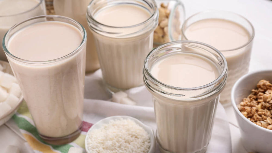 Three glasses of plant-based milk on a white surface with grains.